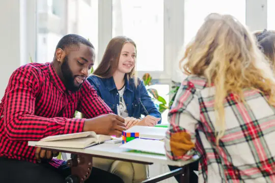 Eine Gruppe von vier Studenten unterschiedlicher ethnischer Herkunft arbeitet gemeinsam an einem Projekt in einem hellen Klassenzimmer. Ein afroamerikanischer Mann im roten Karohemd und eine kaukasische Frau in einer blauen Jeansjacke diskutieren lächelnd über ein Lehrbuch, während andere Studenten neben ihnen ebenfalls engagiert teilnehmen. Diese Szene illustriert die internationale Zusammenarbeit von Studierenden, die möglicherweise mit einem Aufenthaltstitel nach § 16b AufenthG in Deutschland studieren.