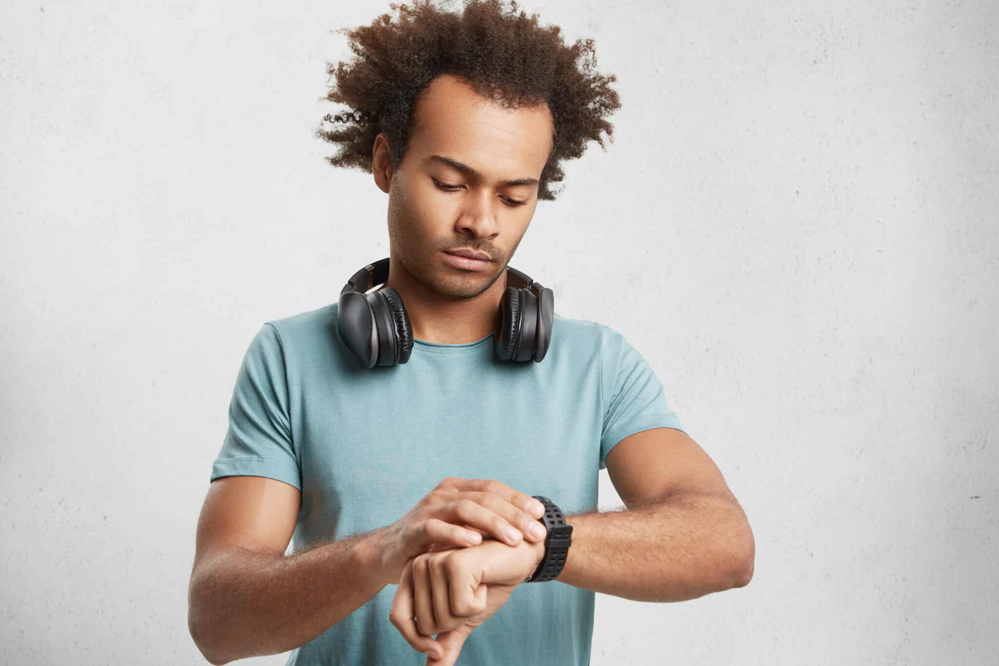 A young man with frizzy hair and headphones around his neck looks at his watch, symbolizing the wait associated with the expiry of the naturalization period. He wears a turquoise T-shirt and stands in front of a neutral white background.
