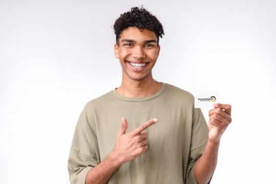 在这张照片中，您可以看到一名非裔美国男子一手拿新居留许可机会卡。他的另一只手正指着卡片。这位年轻人身穿米色 T 恤。