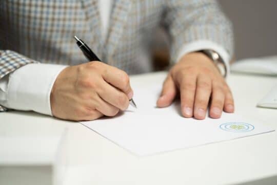 一名身穿白衬衫的男子坐在桌前，在一份盖有公章的官方文件上写字。他正在认真填写表格，可能是根据《居留法》第 16d 条的规定申请居留许可，以便在德国承认外国专业资格。