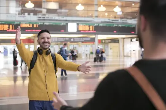 Ein Mann mit Aufenthaltserlaubnis nach § 22 AufenthG freut sich, einen Freund am Bahnhof zu begrüßen. Er trägt ein gelbes Strickoberteil, hat Kopfhörer um den Hals und breitet die Arme für eine Umarmung aus. Im Hintergrund sind Reisende und Anzeigetafeln zu sehen, die die Ankunftszeiten anzeigen