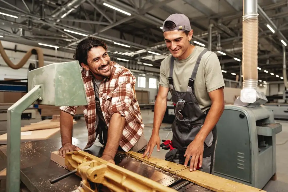 Zwei lachende Tischler, einer mit südeuropäischer und der andere mit osteuropäischer Herkunft, arbeiten zusammen an einer Holzbearbeitungsmaschine in einer gut beleuchteten Werkstatt. Sie tragen Arbeitskleidung und Sicherheitsausrüstung, während Holzspäne um sie herum fliegen, was eine dynamische und freundliche Arbeitsumgebung darstellt