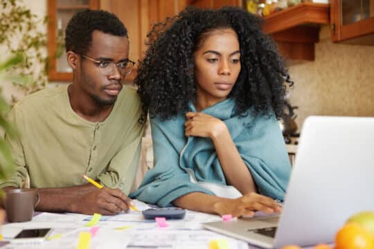 Afroamerički par istražuje dokumente za naturalizaciju kao supružnik u Njemačkoj i saznaje za zahtjeve. Par pogleda u laptop. Čovek hvata beleške na parčetu papira.