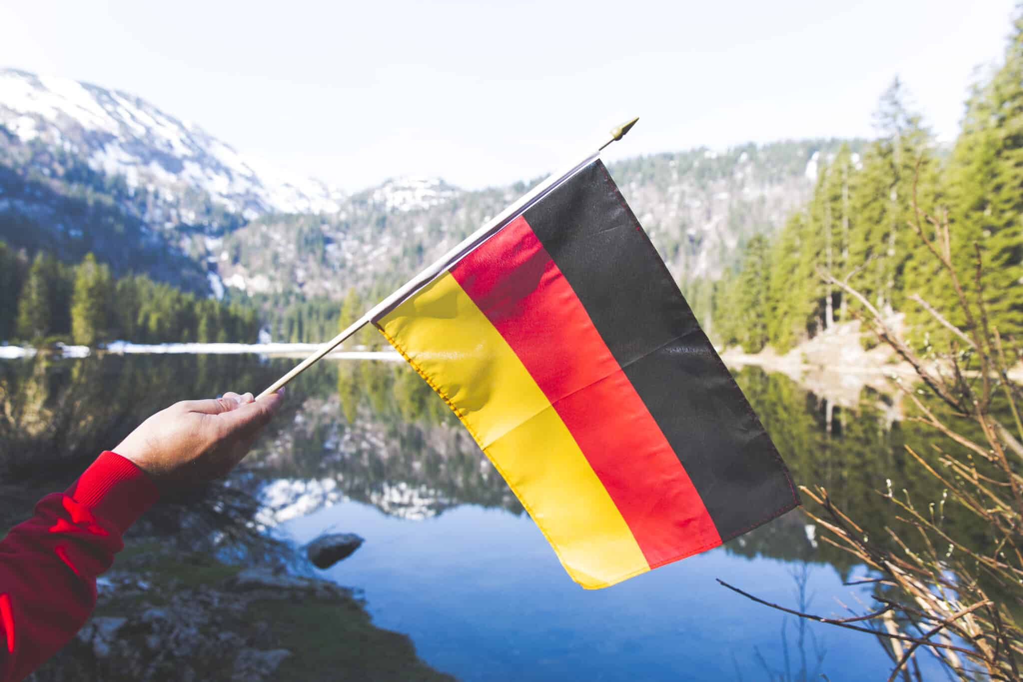 Eine Hand hält eine deutsche Flagge vor einem malerischen See mit Bergen im Hintergrund. Dies symbolisiert das neue Einbürgerungsgesetz 2024, das am 27. Juni 2024 in Kraft tritt und den Weg zur deutschen Staatsbürgerschaft erleichtert.