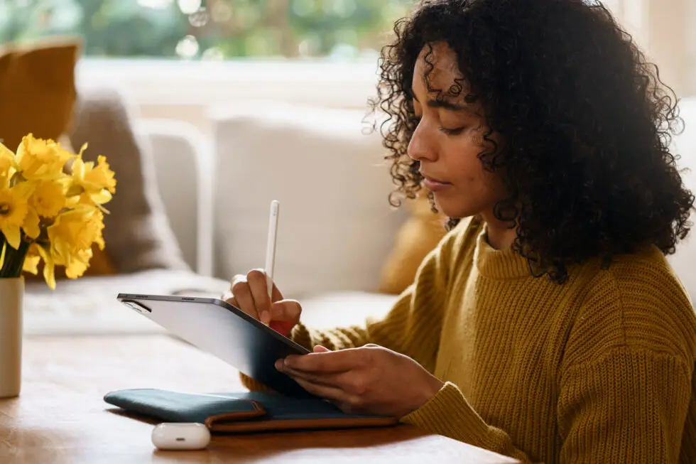 Eine junge Frau sitzt an einem Tisch und nutzt ein digitales Tablet. Sie trägt einen gelben Pullover und ist in eine konzentrierte Tätigkeit vertieft. Auf dem Tisch liegen ein Stift und ein Dokument. Das Bild thematisiert die Vorbereitung und das Sammeln der notwendigen Unterlagen und Informationen zu den Voraussetzungen für die Niederlassungserlaubnis in Deutschland.