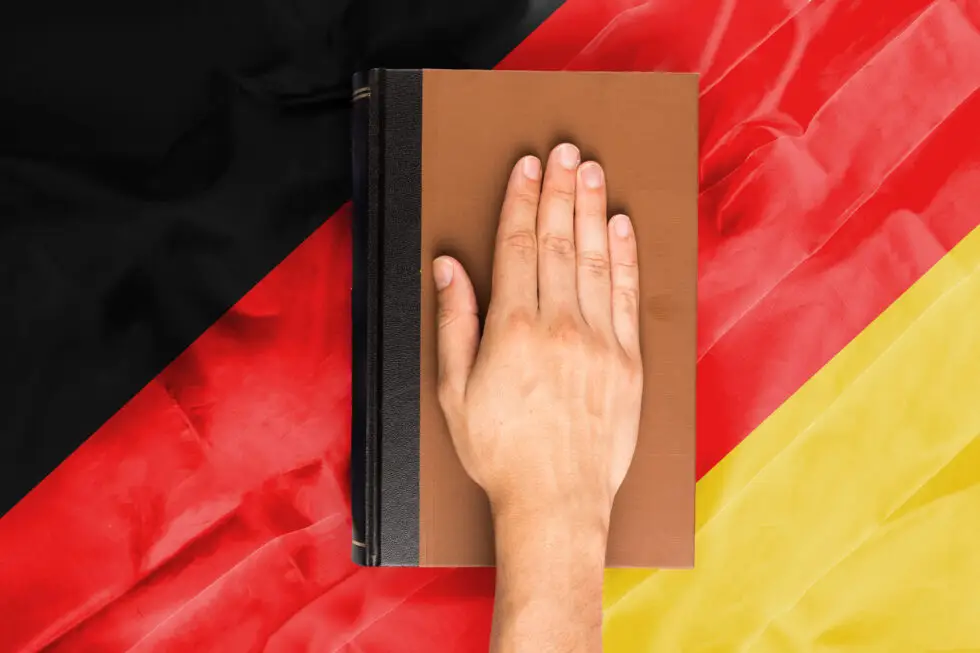 Eine Hand liegt auf einem Verfassungsbuch im Hintergrund sind die schwarz,rot,goldenen Farben Deutschlands. Das Bild symbolisiert die Einbürgerung in Deutschland.