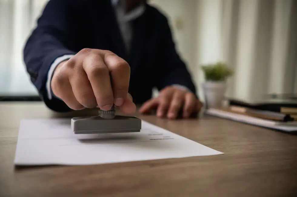 Eine Hand stempelt ein offizielles Dokument auf einem Schreibtisch. Dieses Bild symbolisiert den bürokratischen Prozess und die Genehmigung der notwendigen Unterlagen für die Beantragung einer unbefristeten Aufenthaltserlaubnis ohne B1 Zertifikat.