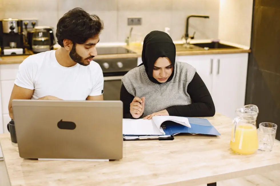 Ein junges Paar, darunter eine Frau mit Hijab, sitzt an einem Küchentisch und arbeitet gemeinsam an einem Laptop und einem Notizbuch. Dieses Bild veranschaulicht das gemeinsame Lernen und die Vorbereitung auf die Beantragung einer unbefristeten Aufenthaltserlaubnis ohne B1 Zertifikat durch alternative Nachweise und Dokumentationen.