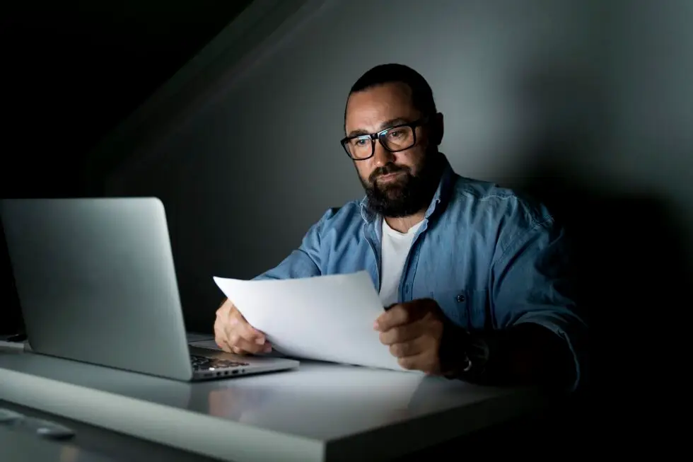 Ein Mann mit Brille arbeitet spät abends am Laptop und hält ein Dokument in der Hand. Er prüft Unterlagen, möglicherweise im Zusammenhang mit seinem unbefristeten Aufenthaltstitel und dem Ablauf seines Reisepasses. Wenn Ihr Aufenthaltstitel unbefristet abgelaufen ist, erfahren Sie hier, wie Sie den Passübertrag durchführen und Ihre Dokumente aktualisieren können.