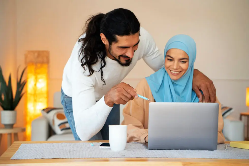 Ein muslimisches Ehepaar guckt gemeinsam auf einen Laptop und lacht. Der Mann steht und deutet mit einem Kugelschreiber auf den Laptop. Seine Frau sitzt am tisch und lächelt. Neben dem Laptop liegen ein Iphone und eine weiße Kaffeetasse.