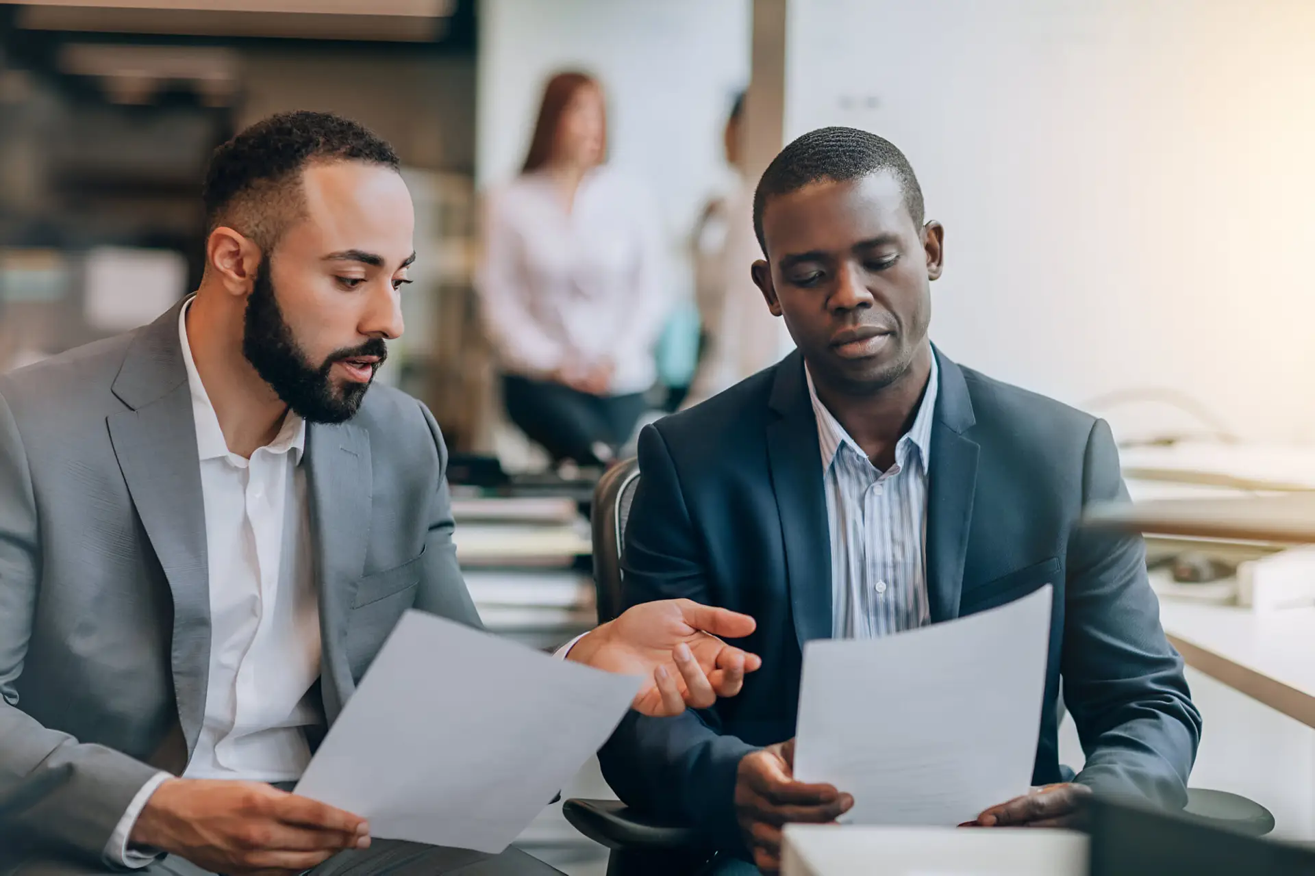 Zwei Männer im Anzug sitzen in einem Büro und besprechen Dokumente. Der eine Mann hält ein Papier in der Hand und erklärt dem anderen Mann die Voraussetzungen für die Niederlassungserlaubnis in Deutschland.