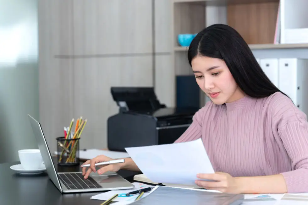 Junge Frau arbeitet konzentriert an ihrem Laptop und prüft Dokumente, während sie sich auf den Prozess vorbereitet, die Deutsche Staatsbürgerschaft zu beantragen. Die Szene zeigt eine moderne Büroumgebung mit einem Drucker im Hintergrund und verschiedenen Büroartikeln auf dem Schreibtisch.