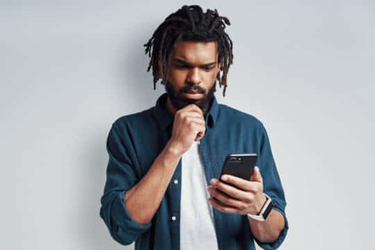 Ein nachdenklicher junger Mann mit Dreadlocks hält ein Smartphone in der Hand und schaut konzentriert auf den Bildschirm. Er informiert sich über das Thema Elektronischer Aufenthaltstitel und welche Voraussetzungen dafür nötig sind. Er trägt ein blaues Hemd über einem weißen T-Shirt.