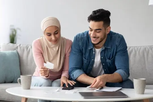 Ein lächelndes Paar, darunter eine Frau mit Hijab, sitzt auf dem Sofa und arbeitet gemeinsam an ihrem Familienbudget. Sie überprüfen Rechnungen und machen Notizen. Das Bild veranschaulicht die sorgfältige Planung und Sicherung des Lebensunterhalts, ein wichtiger Schritt zur Einbürgerung in Deutschland.