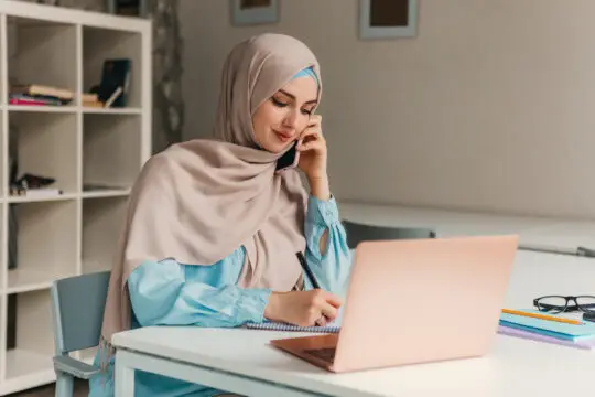 Eine junge Frau mit Hijab sitzt an einem Schreibtisch und arbeitet an ihrem Laptop. Während sie telefoniert, macht sie Notizen in ein Heft. Sie informiert sich über den Antrag auf Bafög, um finanzielle Unterstützung für ihr Studium zu beantragen. Der digitale Antrag erleichtert ihr den Prozess der Beantragung von Bafög