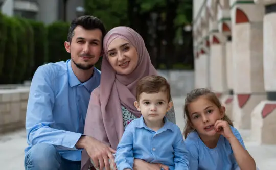 Junge muslimische Familie mit zwei Kindern lächelt im Freien. Die Eltern und Kinder tragen blaue Kleidung, die Mutter trägt ein Kopftuch. Dieses Bild symbolisiert familiären Zusammenhalt, ein zentrales Thema im Kontext des Familiennachzugs nach § 36 AufenthG, insbesondere bei außergewöhnlicher Härte.