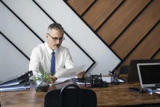 Älterer Mann mit Brille im Büro, der konzentriert Dokumente liest und prüft. Diese Szene veranschaulicht die gründliche Vorbereitung und Überprüfung von Unterlagen, die für die Abgabe einer Verpflichtungserklärung im Aufenthaltsrecht unerlässlich sind.