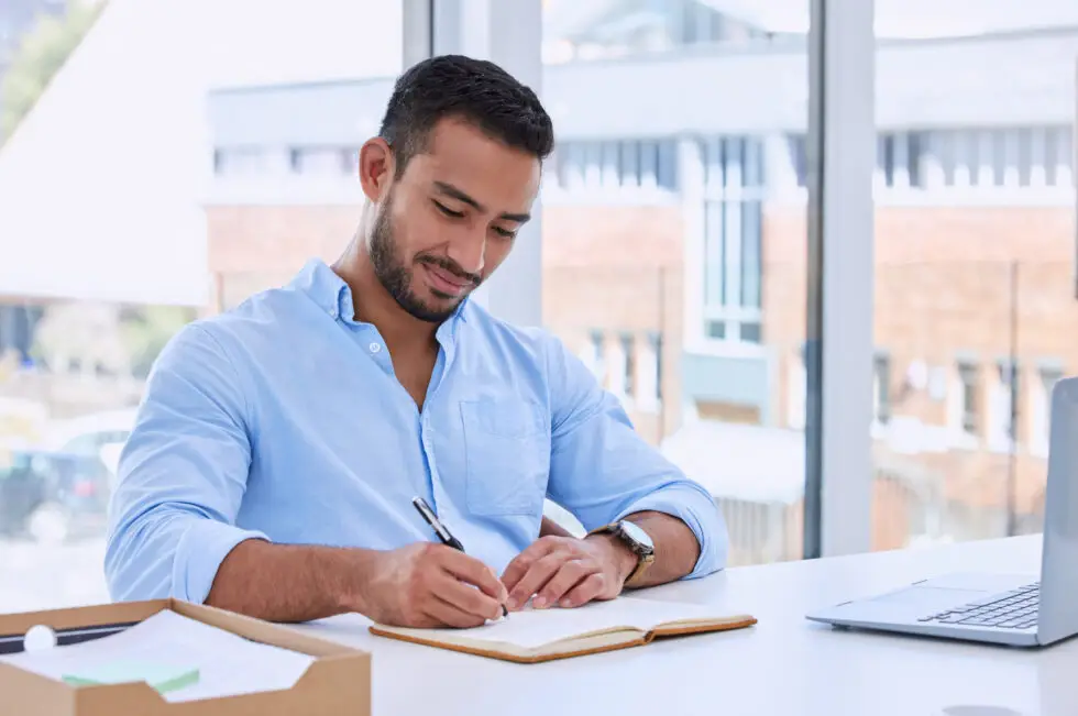 Ein Mann sitzt am Schreibtisch, schreibt konzentriert in ein Notizbuch und plant seinen nächsten Schritt. Dieses Bild symbolisiert die Vorbereitung auf den Antrag des Aufenthaltstitels nach § 16f AufenthG, der Schülern den Schulbesuch oder die Teilnahme an Sprachkursen in Deutschland ermöglicht.