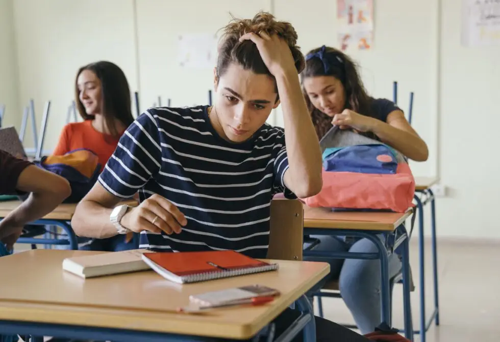 Ein Schüler sitzt nachdenklich und gestresst an seinem Schreibtisch im Klassenzimmer, umgeben von Mitschülern. Diese Szene spiegelt den Druck wider, den viele Schüler beim Erreichen ihres Schulabschlusses erleben, welcher für die Einbürgerung von Bedeutung sein kann. Ein Schulabschluss kann in einigen Fällen den Nachweis von Sprachkenntnissen wie dem B1-Zertifikat ersetzen.