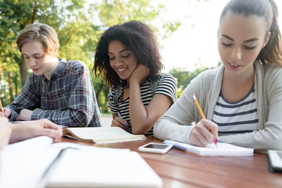 Eine Gruppe internationaler Schüler sitzt im Freien und arbeitet gemeinsam an Studienmaterialien. Dieses Bild symbolisiert den interkulturellen Austausch und den Bildungsweg, der durch den Aufenthaltstitel nach § 16f AufenthG ermöglicht wird, um in Deutschland eine Schule zu besuchen oder einen Sprachkurs zu absolvieren.