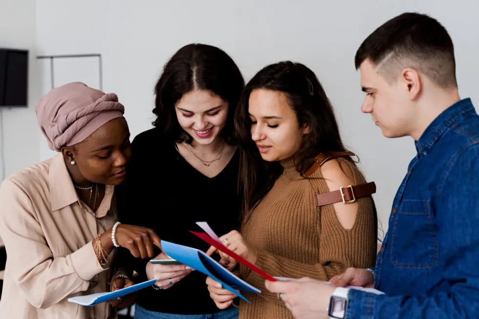 Eine Gruppe internationaler Schüler steht zusammen und diskutiert über Lernunterlagen. Das Bild zeigt den interkulturellen Austausch und die Zusammenarbeit, die durch den Aufenthaltstitel nach § 16f AufenthG ermöglicht wird, der internationalen Schülern den Schulbesuch in Deutschland erleichtert.