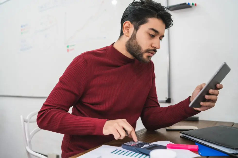 Junger Mann in einem roten Pullover konzentriert bei der Arbeit mit einem digitalen Tablet und Taschenrechner, während er Dokumente prüft. Diese Szene symbolisiert die sorgfältige Vorbereitung und finanzielle Planung, die für die Abgabe einer Verpflichtungserklärung notwendig ist.