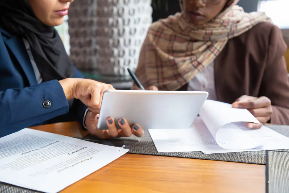 Zwei Frauen, eine mit Kopftuch, arbeiten gemeinsam an Dokumenten und nutzen ein Tablet, während sie rechtliche Informationen zu § 31 AufenthG besprechen, um das Aufenthaltsrecht nach der Trennung zu sichern.