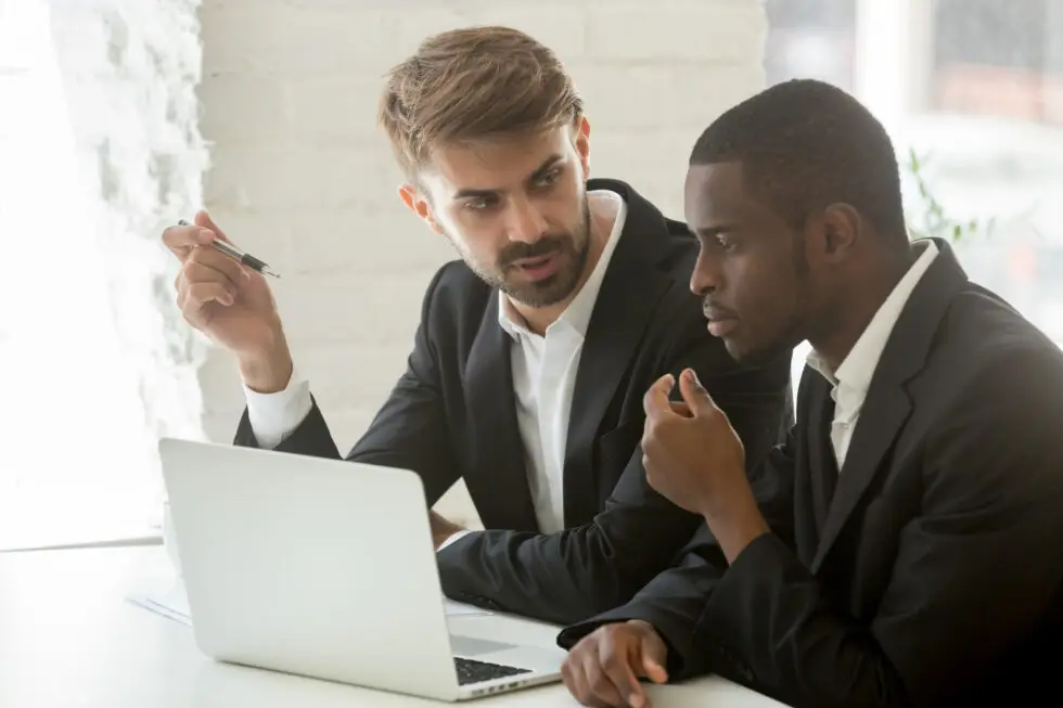 Due uomini d'affari a colloquio davanti a un computer portatile, uno che spiega all'altro informazioni importanti. Questa scena simboleggia la consultazione tra un datore di lavoro e un dipendente straniero in merito all'approvazione dell'impiego ai sensi del § 39 dell'AufenthG, per garantire che tutti i requisiti legali per l'assunzione in Germania siano soddisfatti.