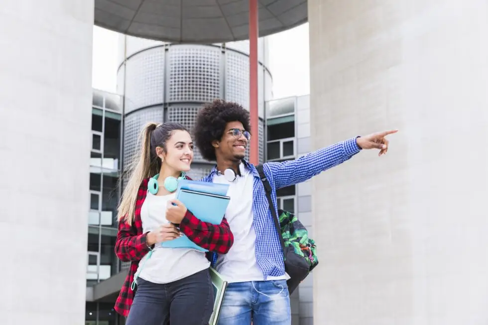 wei internationale Schüler stehen lächelnd vor einem modernen Universitätsgebäude, der männliche Schüler zeigt in die Ferne, während die weibliche Schülerin Bücher hält. Dieses Bild symbolisiert den Weg in eine erfolgreiche Bildungskarriere in Deutschland, der durch den Aufenthaltstitel nach § 16f AufenthG für den Schulbesuch möglich wird.