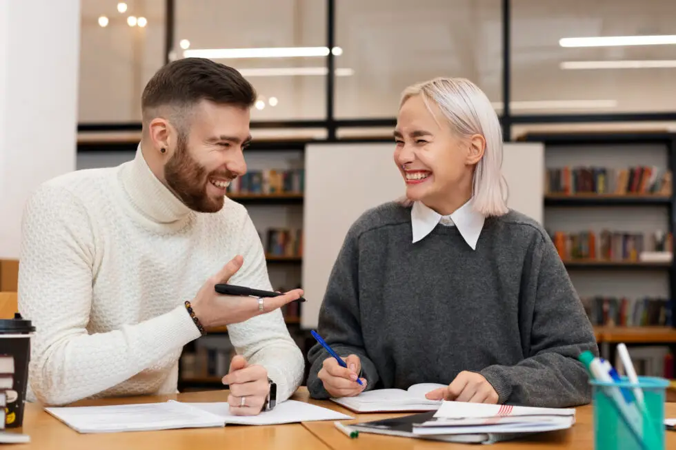 Zwei lachende Personen sitzen an einem Tisch und unterhalten sich während einer Lernsitzung. Das Bild symbolisiert die entspannte Kommunikation und den Austausch einfacher Sprachkenntnisse, wie sie im Alltag erforderlich sind. Es passt zum Thema der Niederlassungserlaubnis, bei der unter bestimmten Umständen einfache mündliche Verständigung ausreicht, um die Sprachvoraussetzungen zu erfüllen.