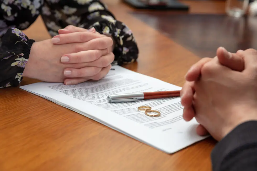 Zwei Personen sitzen sich gegenüber, die Hände gefaltet über einem Trennungsdokument mit Eheringen und einem Stift auf dem Tisch. Das Dokument bezieht sich auf rechtliche Aspekte von § 31 AufenthG zur Sicherung des Aufenthaltsrechts nach einer Scheidung.