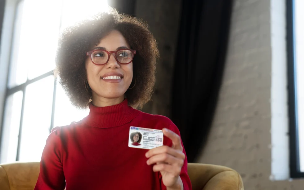 Una mujer sonriente con el pelo rizado y un jersey rojo sostiene una tarjeta que representa su protección subsidiaria en Alemania.